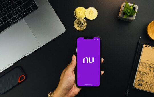 A person holds a smartphone with the Nubank bank app running on the screen. On a table behind the phone, artistic representations of cryptoassets are visible, along with a laptop computer.